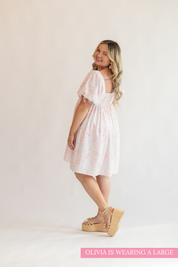 Woman in a white dress with pink floral pattern in front of a white background, looking over her shoulder