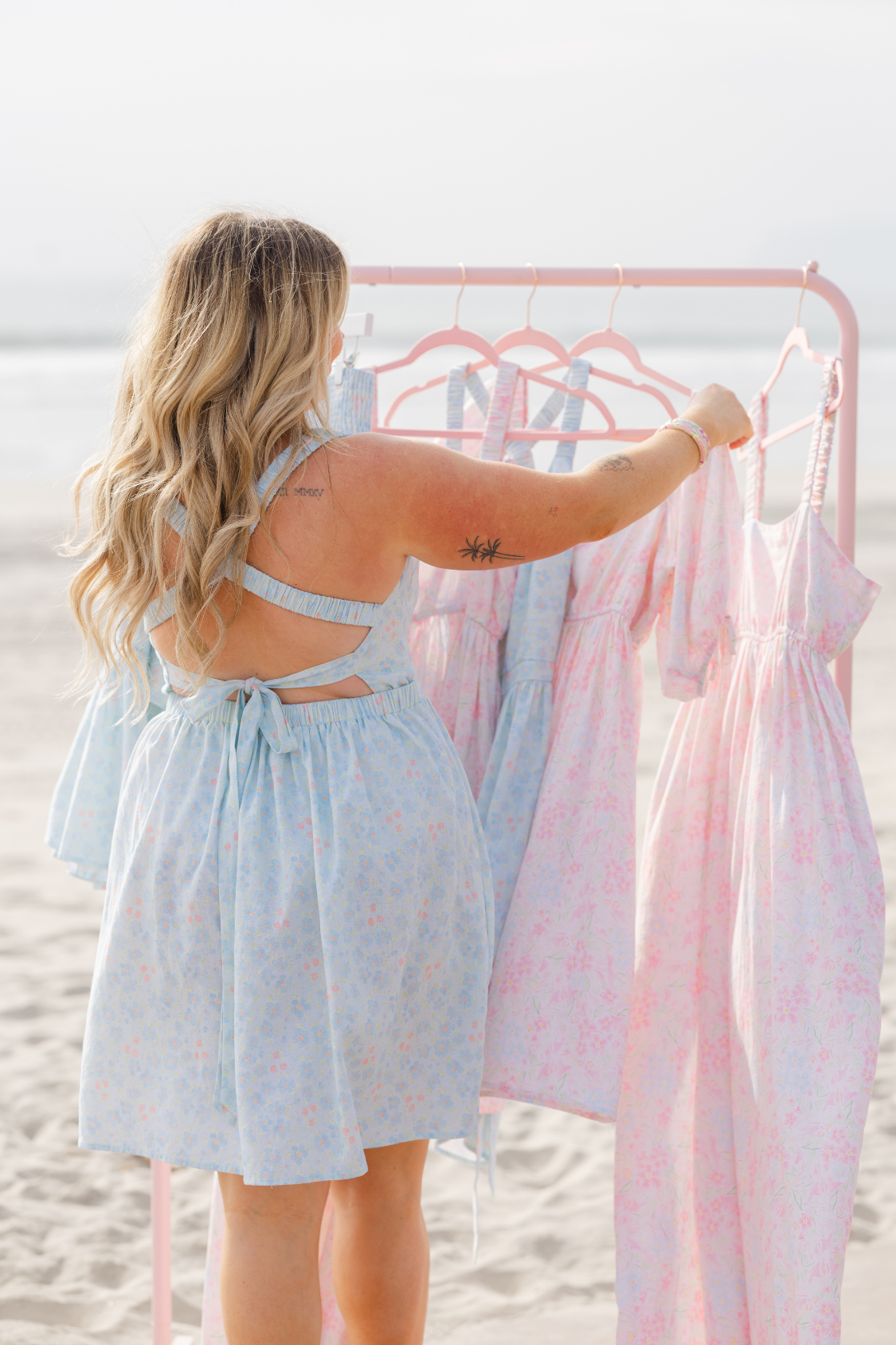 Woman holding a light blue dress with a pink rack of dresses on a beach.