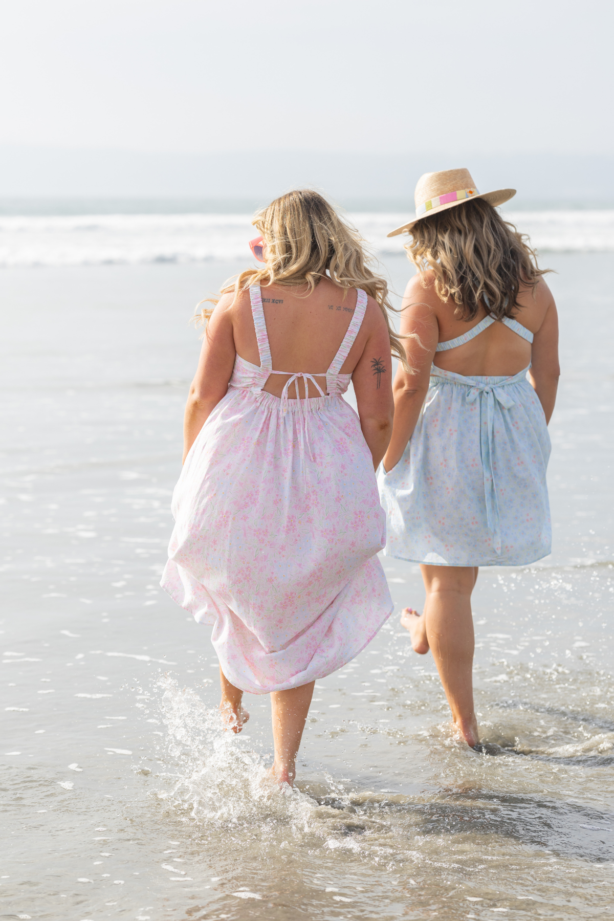 Two women walking on a beach wearing dresses and hats.