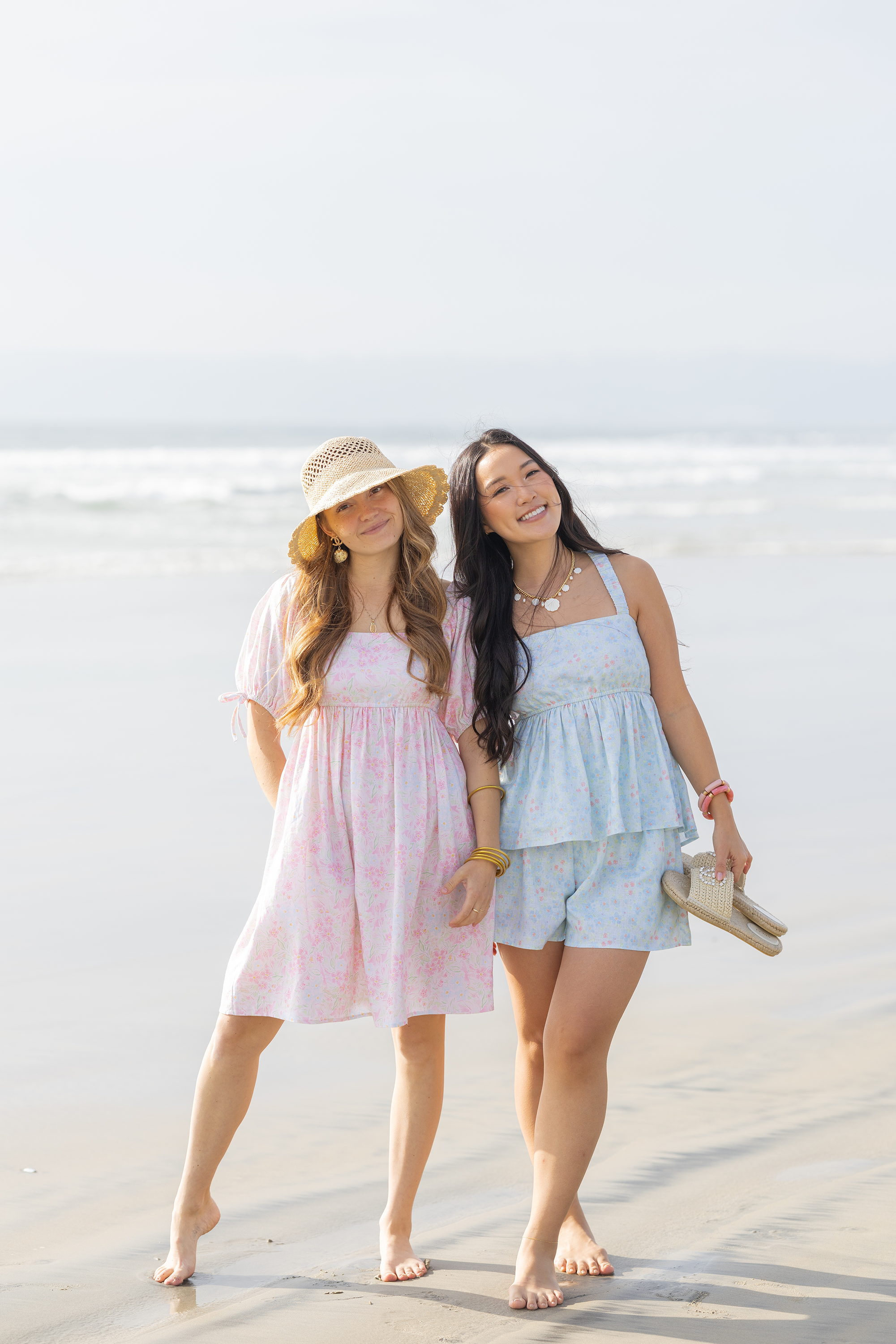 Two women standing on a beach wearing light-colored dresses and hats.