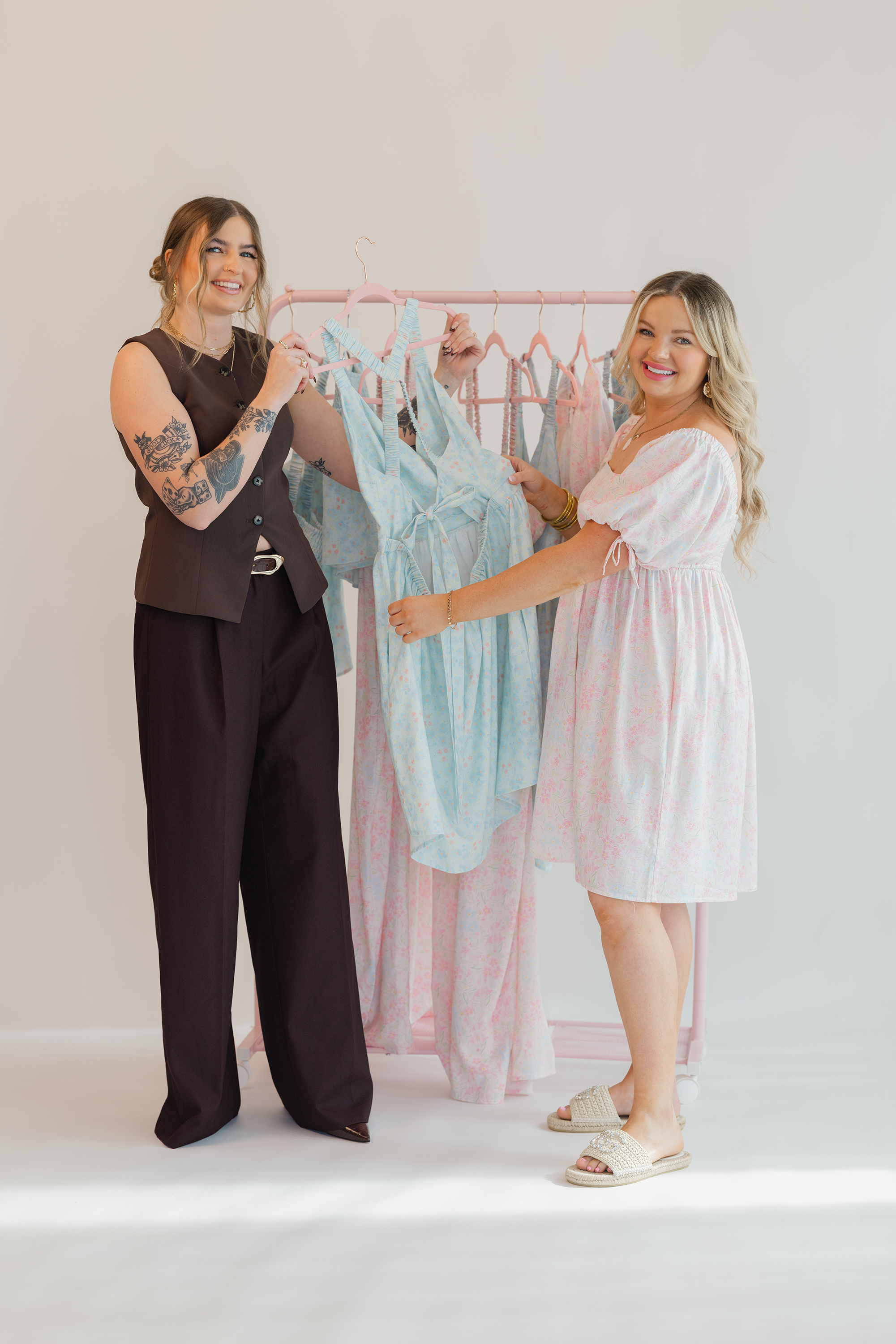 Two women holding a rack of dresses against a plain background